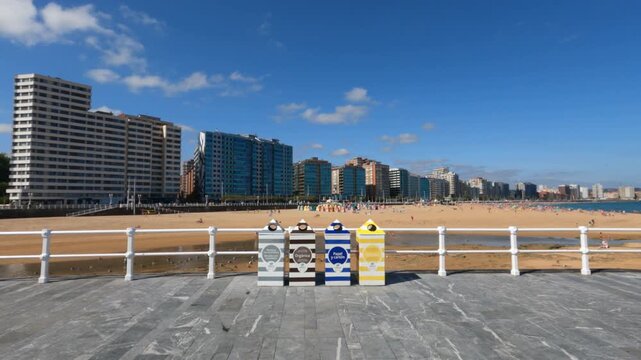 Gijon waterfront skyline and multiple trash cans marking refuse recycling of various types, modern buildings rise beyond a broad seaside promenade