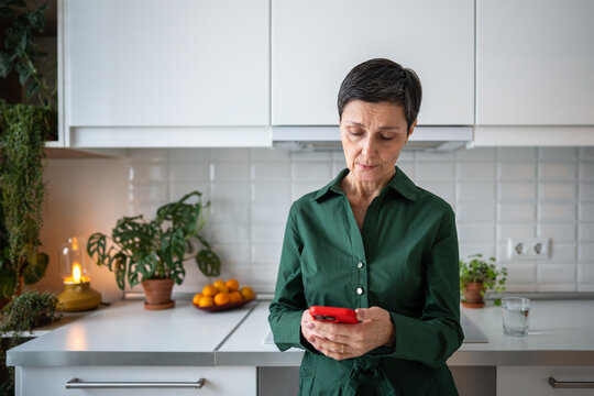 Thoughtful mature woman receiving bad news from smartphone message at home. Serious senior female reading SMS on mobile phone at kitchen, deep in thought, thinking, looking puzzled and worried. 