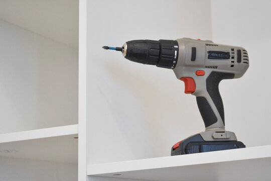 Close-up of a grey and red cordless drill resting on a shelf of a white modular shelving unit indoors