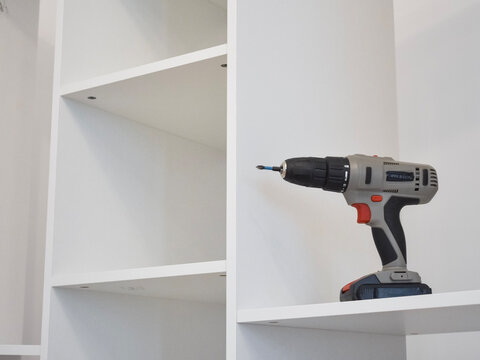 Close-up of a grey and red cordless drill resting on a shelf of a white modular shelving unit indoors