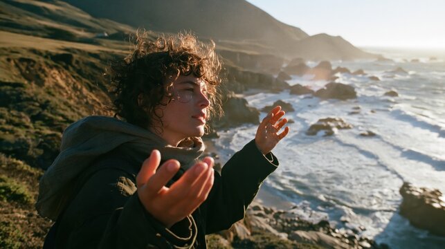 Young man gestures expressively on rugged coastal cliff above sunlit ocean waves