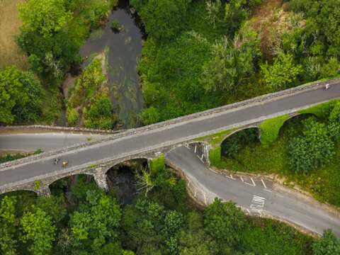 Aerial view of Durrow Viaduct surrounded by lush green trees and vegetation, with cyclists riding across the stone arched bridge over a stream in County Waterford, Ireland.