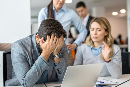 Stressed businessman holding head in hands with worried colleagues supporting him in office