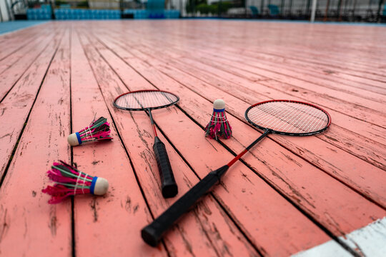 Badminton rackets and shuttlecocks on a weathered pink wooden court surface; sport equipment ready for a game, with a blurred outdoor sports venue background.