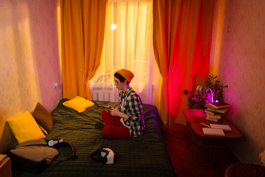 Teenage girl in a beanie using phone while sitting comfortably on bed in softly lit sunset room. Indoor lifestyle and mobile communication at home. Wide shot, top view