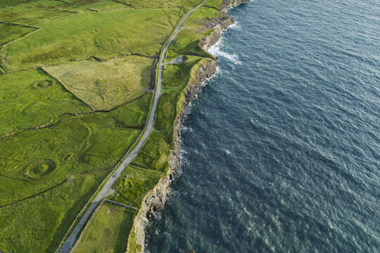 Aerial view of the Cliffs of Moher featuring lush green fields, a winding coastal road, and rugged cliffs meeting the Atlantic Ocean in County Clare, Ireland.