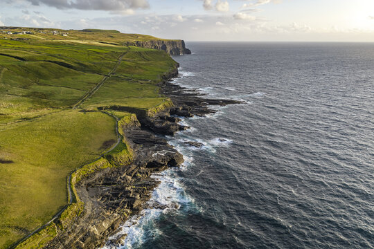 Aerial view of the Cliffs of Moher with rugged green coastal cliffs and waves crashing against the rocks in County Clare, Ireland.