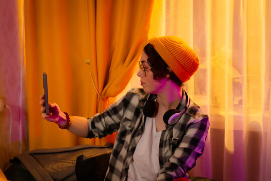 Portrait of teenage girl enjoys free time at home sitting on bed with headphones and beanie while taking smartphone selfie. Room lit by warm sunset light