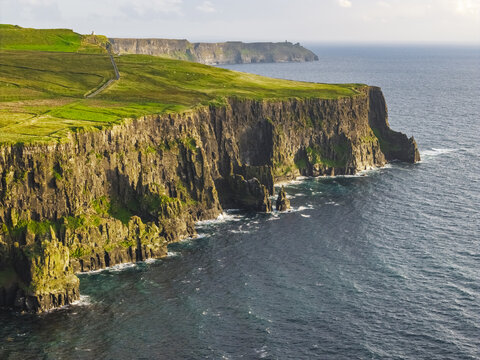 Aerial view of the Cliffs of Moher with lush green grassy fields atop steep rugged rock formations meeting the Atlantic Ocean in County Clare, Ireland.