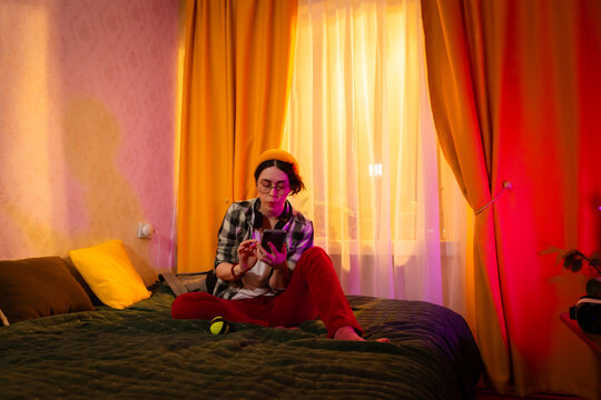 Young girl sitting on bed texting in warmly lit room. Concept of teen routine and digital world. Wide shot