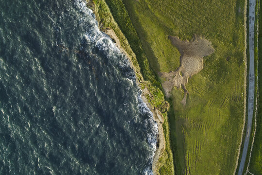 Aerial view of the rugged coastline with crashing waves, green grassy fields, and a coastal road in Doolin, County Clare, Ireland.