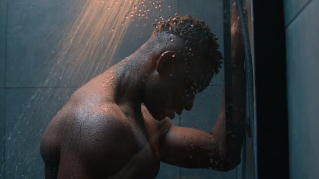 African American muscular man washing wet body under falling shower water in dark bathroom tired exhausted athlete guy male refreshing after workout doing hygiene bodycare cleaning skin indoors bath