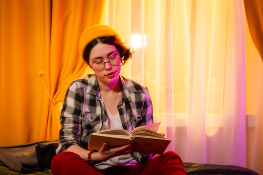 Casual dressed woman sits on bed reading quietly during golden hour. Sunset ambiance, domestic interior, and focus on study and leisure at home. Mid shot