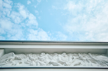 White carved frieze relief showing figures and winged creatures beneath building cornice, captured from low viewpoint with broad pale blue sky and soft clouds above, creating clean historical © Dima Anikin