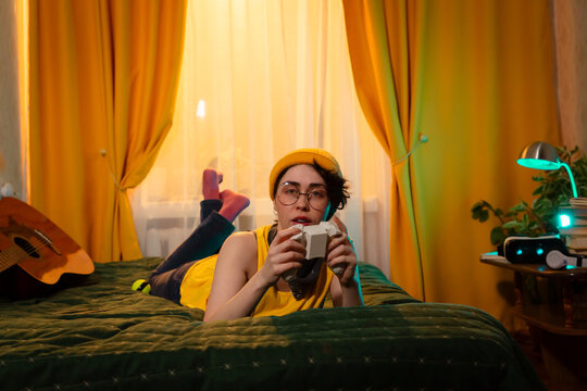 Teenage girl plays console game in a cozy room with retro lamp and pillows. Concept of home comfort and youth habits. Mid shot