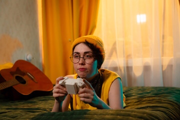 Teen gamer girl in colorful outfit and beanie hat lies on bed with a gamepad. Concept of relaxed habits and teenage personality. Mid shot © _KUBE_