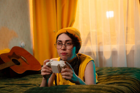 Teen gamer girl in colorful outfit and beanie hat lies on bed with a gamepad. Concept of relaxed habits and teenage personality. Mid shot