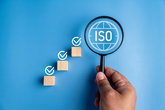Hand holding magnifying glass over ISO standard symbol with ascending wooden steps of checkmarks on blue background for quality audit
