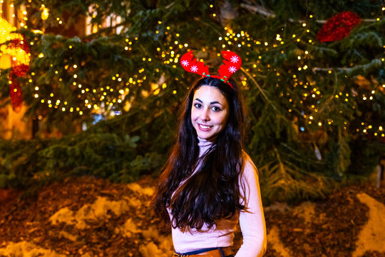 Young woman smiling with reindeer antlers headband in front of holiday tree lights