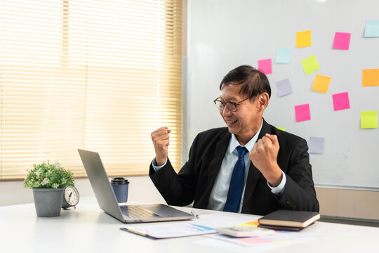 Happy senior asian businessman celebrating business success with raised fists beside laptop in modern office
