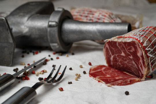 Dry-cured pork coppa wrapped in netting with a slice next to a vintage metal meat grinder and peppercorns.