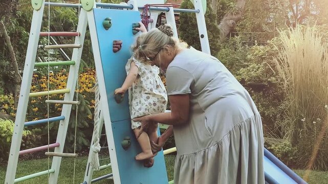 Grandmother helps a toddler girl climb an outdoor play structure, supportive grandparenting, active childhood, family bonding and summer play.