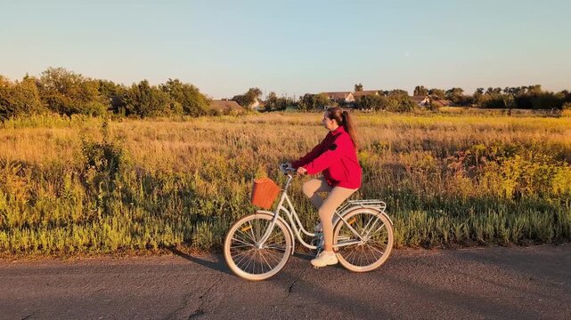 A girl rides her bike along a path next to a large field during sunset. The sun lights up the grass and trees around her