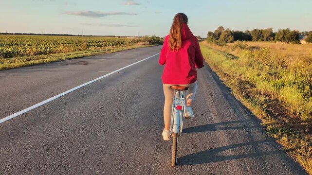 A person rides a bicycle down a long road while fields are on either side. The sun is setting, casting a warm light over the scene