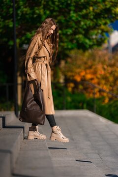 woman in long beige trench coat walking outdoors in urban setting with casual and stylish appearance, side view of a young woman in autumn foliage, natural light, clean background.