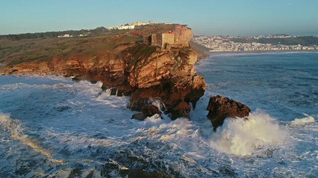 Aerial view of the historic Fort of Sao Miguel Arcanjo perched on a rocky cliff with waves crashing against the shore during golden hour in Nazare, Leiria District, Portugal.