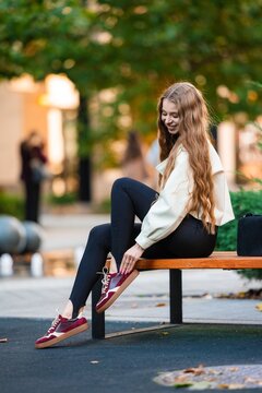 Woman sitting on bench, in outdoor park, with bench, handbag, from a side view perspective, with beige, navy blue accents, and relaxed mood, with nearby objects.