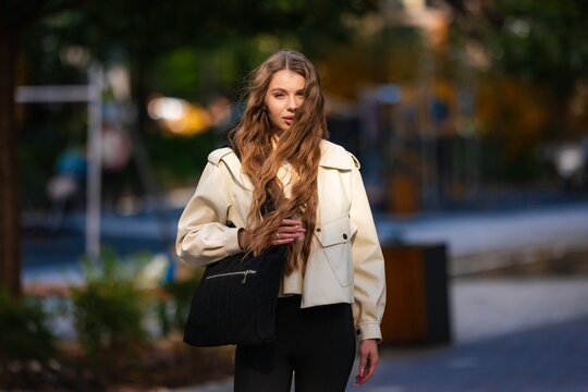 woman walking in urban park with black bag and white jacket, casual front view, beige pants, fashion lifestyle, natural light, clean background, copy space, modern design, vivid colors, web.
