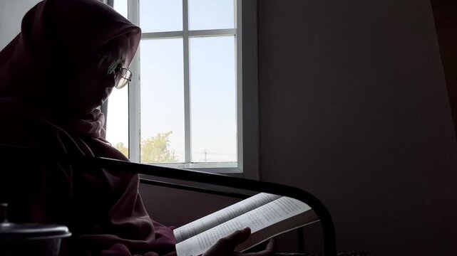 Slow motion low angle side view of an Asian Muslim woman in a hijab and glasses. She is sitting indoors reading a book by a bright window, enjoying a quiet and peaceful study time at home.