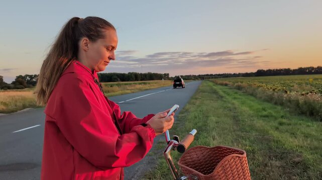 A young woman stands by her bicycle on the roadside while looking at her smartphone. The sun sets behind her in the background