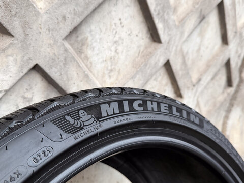 Black rubber sidewall of premium car tire from famous French manufacturer Michelin close up, wheel part with corporate brand logo text and mascot character near decorative concrete wall background.
