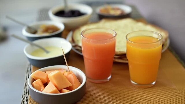 Two glasses of fresh orange and grapefruit juice stand in the foreground of a breakfast spread. Behind them lie thin pancakes, bowls of poppy seed filling, fruit jam, and fruit.