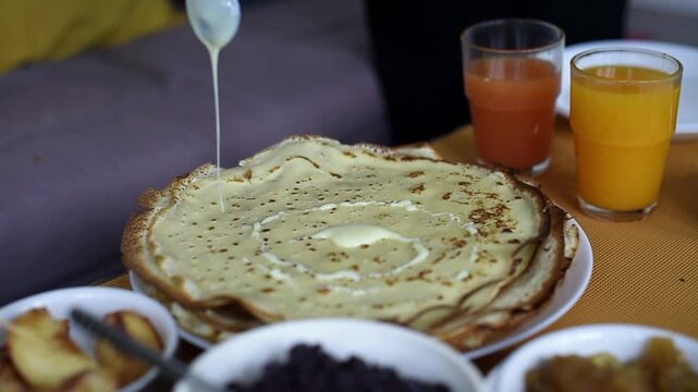 A stream of sweet condensed milk pours from a spoon onto a golden pancake on a breakfast plate. Bowls of poppy seed filling, fruit, and glasses of fresh citrus juice sit nearby.