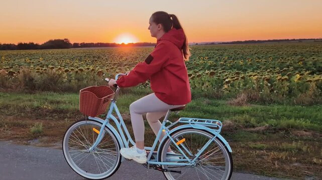 A woman rides a bicycle on a path beside a sunflower field during sunset. The sky shows shades of orange and pink as the sun sets in the distance