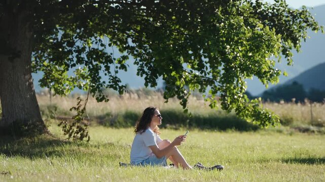 Young woman using a smartphone while sitting on the grass under a large tree, enjoying a peaceful outdoor moment in nature.