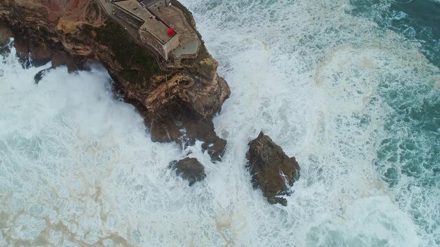 Aerial view of the historic Farol da Nazare lighthouse perched on a rocky cliff with powerful ocean waves crashing against the shore in Nazare, Leiria District, Portugal.