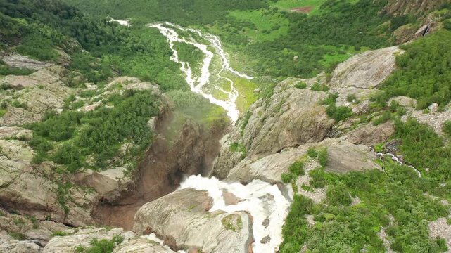 Shdugra Waterfall flows in aerial view from a high rocky ledge toward green alpine slopes below Ushba glaciers, supporting trekking routes, watershed research and Svaneti destination marketing.
