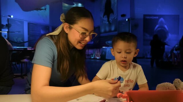 A young Latina mother and her two-year-old son coloring shark-themed pages with crayons in an underwater-themed area, illuminated by blue LED lights at Chapultepec Zoo.