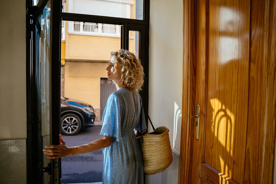 Mature Blonde Woman Entering Apartment Building with Straw Bag on Sunny Day in Spain
