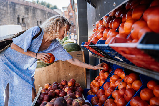 Smiling Mature Blonde Woman Choosing Fresh Tomatoes at Outdoor Produce Market in Spain