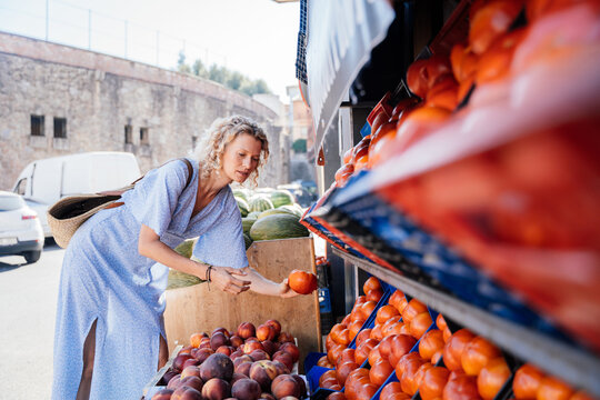 Mature Blonde Woman Holding Ripe Tomato While Shopping Fresh Produce at Spanish Market