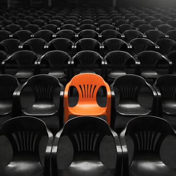 One Bright Orange Chair Standing Out in a Grid of Empty Black Plastic Chairs Concept