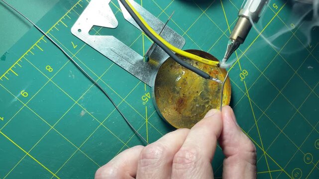 Close-up view of a technician's hand holding a wire while applying a soldering iron to a circular metal base, surrounded by tools and a green cutting mat in a well-lit workspace. High quality 4k