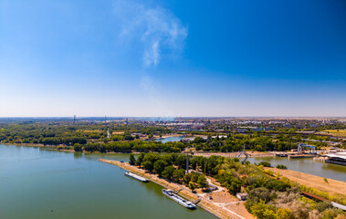Fototapeta premium View of a river landscape and city with clear skies during the day. A river flows through a green landscape with buildings in the distance under a bright blue sky.