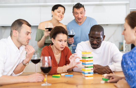 Woman spending free time with group of mates at laid-back home get-together with drinks, concentrating on removing block from tower while playing jenga