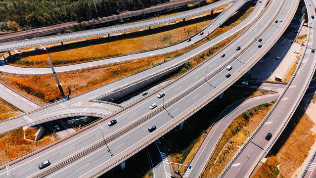 Traffic flows on a busy highway system with multiple lanes and intersections. Cars move along a complex network of highways in a city during the day. Trees line the sides of the roads.